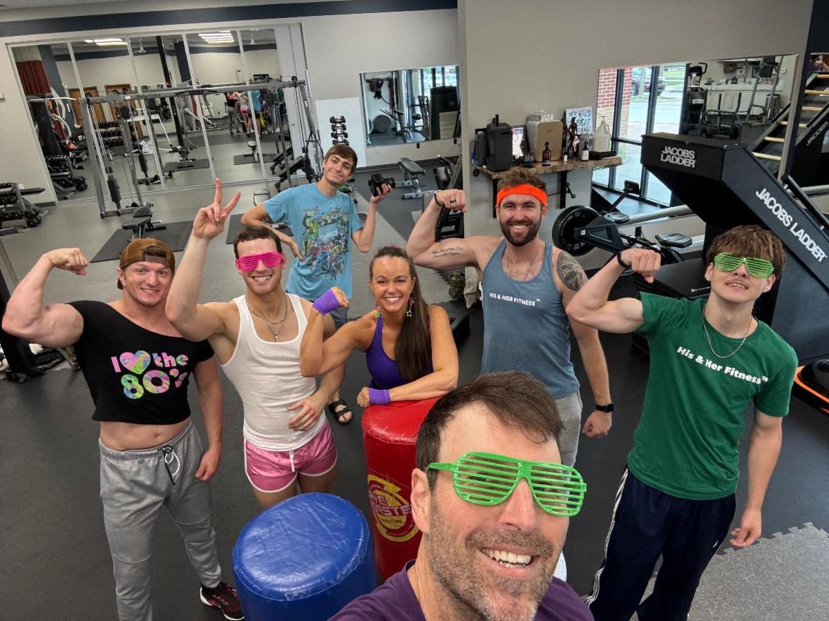 A group in 80s outfits flexing and smiling on the His & Her Fitness studio floor